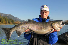 Ein Angler hält einen großen frisch gefangenen Fisch am Flussufer, umgeben von einer malerischen Berg- und Waldlandschaft.
