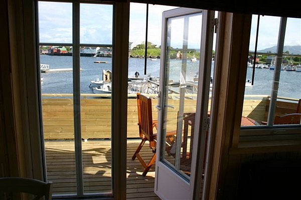 Blick durch geöffnete Terrassentür auf Holzbalkon mit Stühlen und malerischem Fjord in Nordland, Norwegen. Ideal für Angelreisen und Naturerlebnisse.