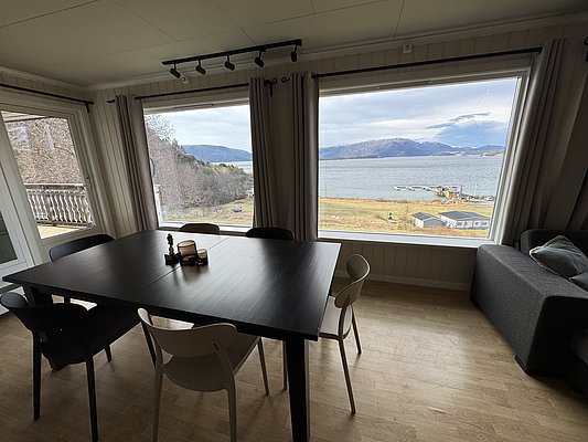 Modernes Wohnzimmer mit großem Esstisch, Panoramafenster bieten atemberaubenden Blick auf norwegische Fjordlandschaft in Hitra. Gemütliche Atmosphäre, perfekt für entspannten Angelurlaub oder Familienausflüge. Fantastische Naturkulisse lädt zum Entdecken ein.