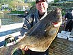 Ein Angler präsentiert stolz einen großen Heilbutt auf einem Steg in Hitra, Norwegen. Die idyllische Umgebung unterstreicht das ideale Angelziel für Meeresangeln und Abenteuerlust. Perfekt für den nächsten Angelurlaub.