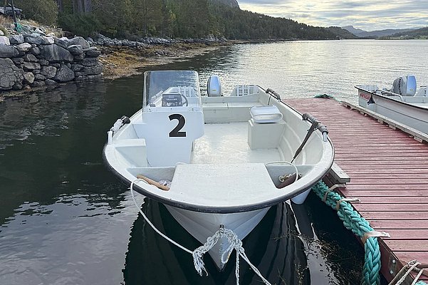 Ein weißes Angelboot liegt an einem Holzsteg in der malerischen Küstenregion Hitra, Norwegen. Ruhiges Wasser und bewaldete Hügel im Hintergrund schaffen eine entspannte Atmosphäre für einen Angelurlaub.
