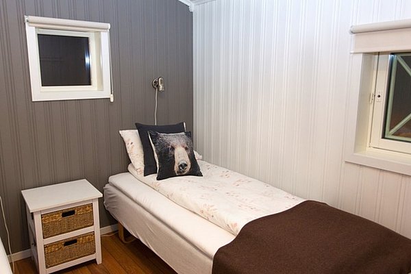 Gemütliches Schlafzimmer in Nord-Norwegen mit Einzelbett, brauner Decke und Kissen mit Tiermotiv. Weiße Holzwände und Fenster sorgen für eine helle, freundliche Atmosphäre. Perfekt für erholsame Angelurlaube in Skandinavien.