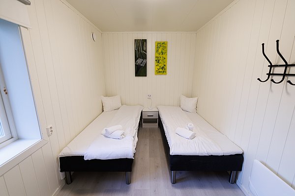 Kompaktes, gemütliches Schlafzimmer in Nordland, Norwegen. Zwei Einzelbetten, minimalistische Einrichtung mit hellen Holzwänden, Kunst an der Wand, ideal für erholsame Nächte nach einem aufregenden Angeltrip in der malerischen norwegischen Landschaft.