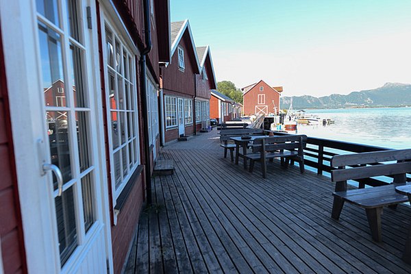 Holzdeck mit Sitzbänken vor roten Fischerhütten in Nordland, Norwegen. Blick aufs Wasser mit Bergen im Hintergrund. Ideal für Angelreisen und Naturerlebnisse.