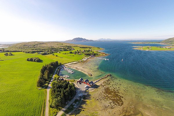 Luftaufnahme aus Nordland, Norwegen: malerische Küstenlandschaft mit grünen Feldern, blauem Meer und typisch roten Häusern. Ideal für Angelreisen und Naturerlebnisse. Perfekt zur Entspannung und Erkundung der beeindruckenden norwegischen Landschaft.