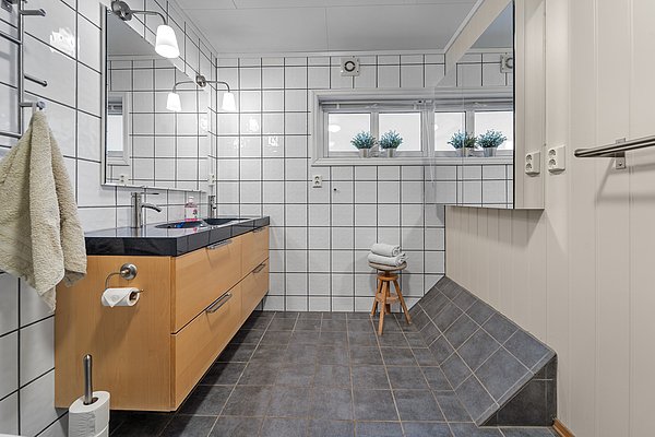 Modernes Badezimmer in Nord-Norwegen mit großem Spiegel, Holzkommode und eleganten Fliesen. Helle Beleuchtung, Fenster mit Pflanzen, und ein Hocker mit Handtüchern sorgen für Komfort und Stil. Ideal für erholsamen Angelurlaub.