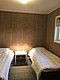 Gemütliches Schlafzimmer in einer Holzhütte auf Hitra, Norwegen. Helle Deckenbeleuchtung, zwei Einzelbetten, Holzvertäfelung und Steinboden schaffen eine einladende Atmosphäre. Ideal für Angelreisende zur Entspannung nach einem erfolgreichen Tag auf See.