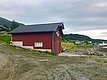 Rotes Holzhaus am Ufer in Nord-Norwegen, umgeben von grünen Hügeln und grauem Himmel. Perfekter Ausgangspunkt für Angelreisen und Naturerlebnisse in malerischer Umgebung. Optimaler Mix aus Tradition und Abenteuer.