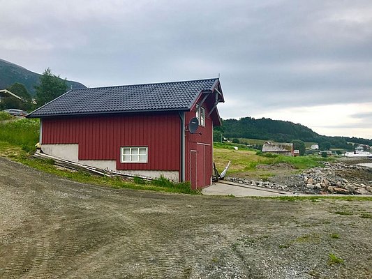 Rotes Holzhaus am Ufer in Nord-Norwegen, umgeben von grünen Hügeln und grauem Himmel. Perfekter Ausgangspunkt für Angelreisen und Naturerlebnisse in malerischer Umgebung. Optimaler Mix aus Tradition und Abenteuer.