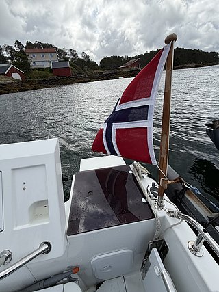 Boot in Süd-Norwegen mit norwegischer Flagge, nahe Küste mit roten Holzhäusern und bewölktem Himmel. Perfekter Ausgangspunkt für erlebnisreiche Angelreisen und Meeresabenteuer.