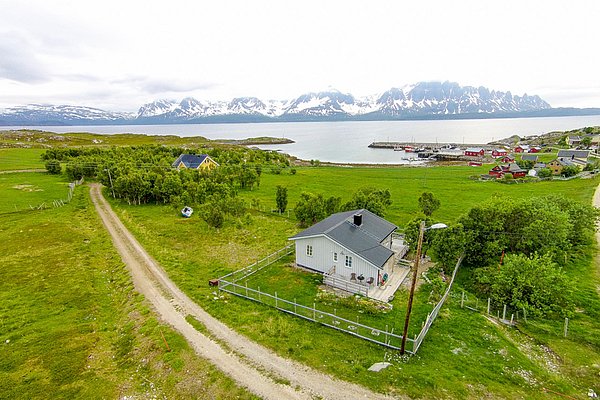 Luftaufnahme einer idyllischen Landschaft in Nord-Norwegen mit kleinen Häuschen, grünen Wiesen und schneebedeckten Bergen im Hintergrund am ruhigen Fjord. Perfekter Ausgangspunkt für Angelurlaub und Outdoor-Abenteuer.