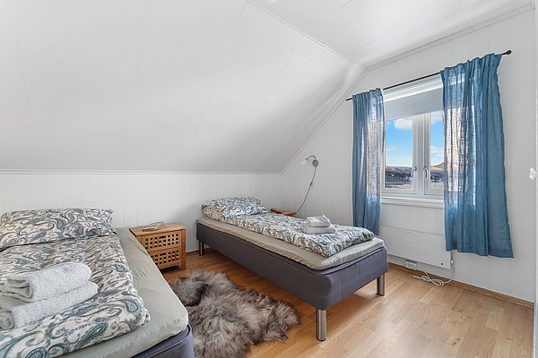 Gemütliches Schlafzimmer in Nord-Norwegen mit zwei Einzelbetten, hellem Holzfußboden und blauen Vorhängen. Fensterblick auf verschneite Berge. Ideal für entspannte Angelreisen und erholsame Nächte nach einem erfolgreichen Angeltag.