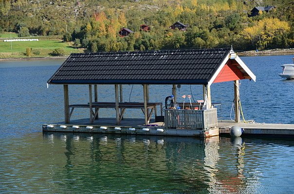 Nord-Norwegen: Malerischer Bootssteg mit überdachtem Bereich auf ruhigem Gewässer, umgeben von herbstlich gefärbten Bäumen. Perfekter Ort für Angelreisende, umgeben von idyllischer Naturkulisse, lädt zum Verweilen und Fischen ein.