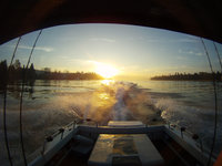Blick von einem Boot auf ein glitzerndes Gewässer mit Sonnenuntergang am Horizont, der die Silhouetten der umgebenden Landschaft hervorhebt.