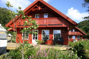 Rotes Holzhaus in Süd-Norwegen, umgeben von grüner Natur. Große Fenster, Balkon und Terrasse bieten gemütlichen Komfort. Perfekt für Angelurlauber, die skandinavische Idylle und erholsame Tage am Wasser suchen. Ideal für naturnahe Erholung.