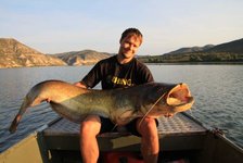 Ein Mann sitzt in einem Boot auf einem großen See, hält einen großen Wels, umgeben von einer malerischen Berglandschaft im Hintergrund.
