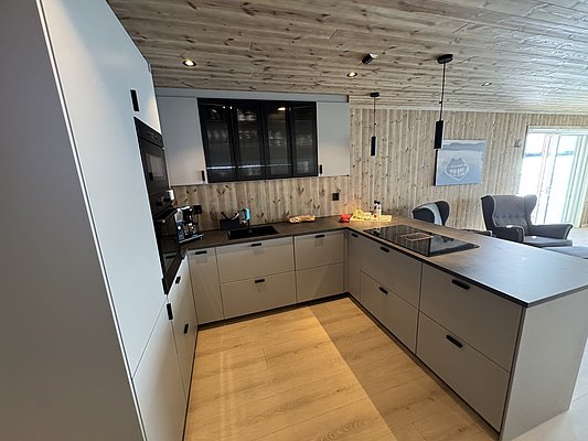 Moderne, offene Küche in Hitra, Norwegen, mit stilvollen grauen Schränken, integrierten Geräten und Holzverkleidung, ideal für Angelurlauber. Helle, einladende Atmosphäre durch große Fenster. Perfekt für Erholung nach einem erfolgreichen Angeltag.