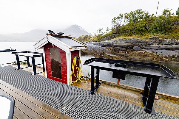 Nordland, Norwegen: Ein malerischer Fischereistand mit roten Schuppen, praktischen Arbeitstischen am Wasser und nebligen Bergen im Hintergrund. Perfekt für Angler, die unberührte Natur und Abenteuer suchen.