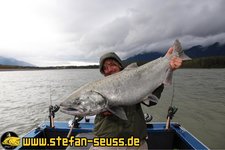 Eine Person steht auf einem Boot auf einem Fluss und hält einen großen Lachs in die Kamera, während Angeln im Hintergrund sichtbar sind.