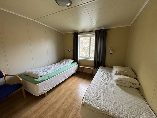 Gemütliches Zimmer in Hitra, Norwegen, mit zwei Einzelbetten, Holzboden und großem Fenster. Ideal für Angelurlauber, die Ruhe und Natur genießen möchten. Angenehme Beleuchtung, schlichtes Design für erholsame Nächte nach erfolgreichen Fischen.