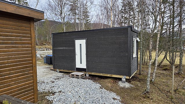 Kleine, moderne Holzhütte in Nordland, Norwegen, umgeben von lichten Bäumen und Schotterweg. Ideal für Angelurlaub und Naturerlebnis. Ruhige, abgelegene Lage in der skandinavischen Wildnis.