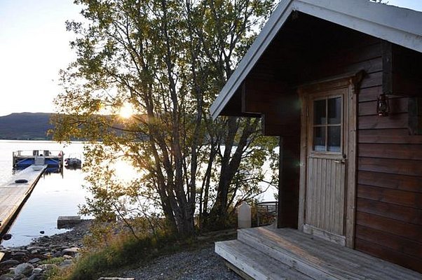 Holzhütte am ruhigen Wasser in Nord-Norwegen. Sonnenuntergang hinter Bäumen, Steg mit Booten. Perfekter Angelurlaubsort für Entspannung und Abenteuer in der Natur. Entdecke fischreiche Gewässer und unberührte Schönheit.