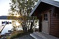 Holzhütte am ruhigen Wasser in Nord-Norwegen. Sonnenuntergang hinter Bäumen, Steg mit Booten. Perfekter Angelurlaubsort für Entspannung und Abenteuer in der Natur. Entdecke fischreiche Gewässer und unberührte Schönheit.