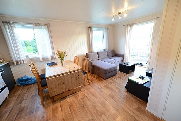 Gemütliches Wohnzimmer in Nord-Norwegen: Holzboden, graues Ecksofa, schlichter Esstisch mit Blumen, viel Tageslicht durch große Fenster. Ideal für entspannte Angelurlaube und naturnahe Erholung.