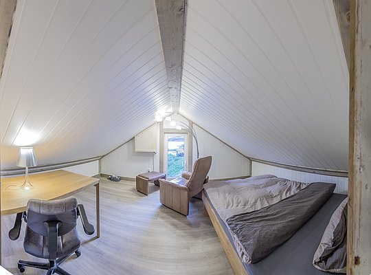 Gemütliches Dachgeschosszimmer in Nordland, Norwegen, mit Holzboden, Doppelbett, Schreibtisch und Sessel. Ideal für Angelurlaub, bietet es Entspannung nach einem erfolgreichen Angelausflug. Helle, freundliche Atmosphäre mit kleinem Fensterblick ins Grüne.