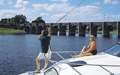 Ein Mann angelt von einem Boot aus, während eine Frau daneben sitzt, mit einer steinernen Bogenbrücke und Flusslandschaft im Hintergrund.