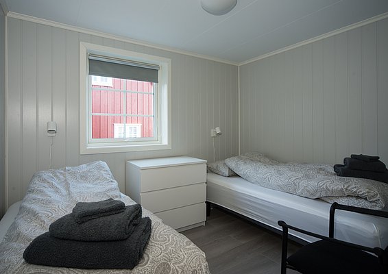Gemütliches Schlafzimmer in Hitra, Norwegen mit zwei Einzelbetten, schlichter Einrichtung und hellen Wänden. Perfekte Unterkunft für Angelreisen, bietet Entspannung nach einem erlebnisreichen Tag. Fenster mit Ausblick auf rotes Holzhaus.