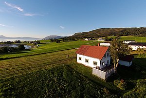 Ländliche Idylle in Nord-Norwegen: Ein weißes Haus mit rotem Dach inmitten grüner Felder, umgeben von Bergen und Meer. Perfekt für Angelurlaub und Erholung in atemberaubender Natur.