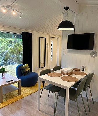 Gemütlicher Wohnbereich in Hitra, Norwegen: Modernes Interieur mit Esstisch, TV und blauem Sessel. Große Fenster bieten Ausblick auf die Natur. Ideal für entspannte Angelurlaube.