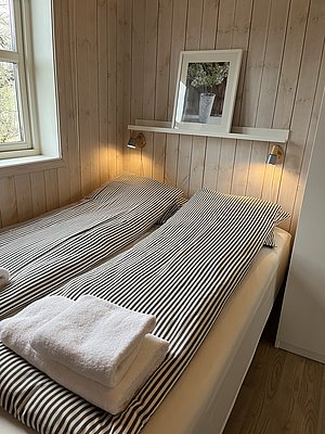 Gemütliches Schlafzimmer mit gestreifter Bettwäsche und Holzverkleidung in der Region Hitra, Norwegen. Warmes Licht und ein Fenster mit Blick ins Grüne schaffen eine einladende Atmosphäre. Ideal für erholsame Angelurlaube.
