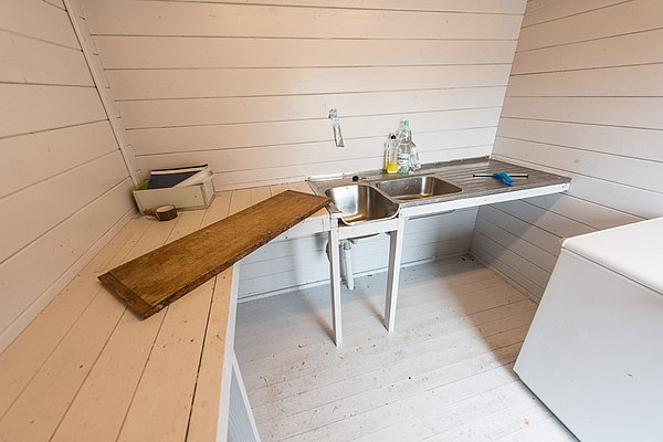 Heller, rustikaler Raum in Nordland, Norwegen. Weißes Holzinterieur, Spüle mit Doppelbecken und Ablagefläche. Ideal für Reinigungsarbeiten nach dem Angeln. Einfacher, funktionaler Stil, typisch für norwegische Fischerhütten.