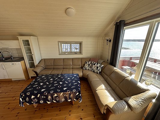 Gemütliches Wohnzimmer in einem Ferienhaus auf Hitra, Norwegen. Helle Holzwände, bequeme Ledersofas, geschmackvolle maritime Dekoration und Meerblick durch große Fenster schaffen eine einladende Atmosphäre für Angler und Naturliebhaber. Ideal für Angelurlaub genießen.