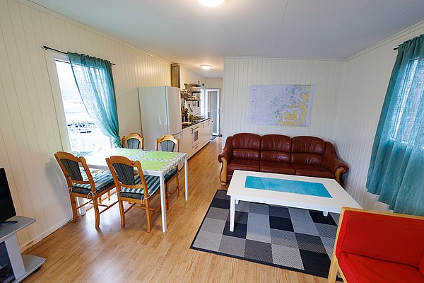 Gemütlicher Wohnraum in Nord-Norwegen: Helle Holzwände, Esstisch, rote Ledercouch, Fenster mit türkisfarbenen Vorhängen. Ideal für entspannte Angelurlaube, nahe atemberaubender Natur und Fischgründen. Perfekt für erholsame Momente nach dem Angeln.