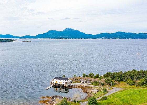 Luftaufnahme von Hitra, Norwegen: Küstenhaus am Wasser, umgeben von üppiger Natur und Bergen im Hintergrund. Ideal für Angelreisen und naturverbundenen Urlaub. Spektakuläre Landschaft zum Entspannen und Entdecken. Perfekter Rückzugsort für Abenteuer und Erholung.
