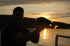 Eine Person hält bei Sonnenuntergang einen Fisch vor einem See, wobei der Fisch durch das Licht hervorgehoben wird.