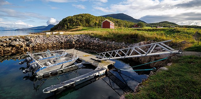 Nord-Norwegen: Malerische Küstenlandschaft mit klaren Gewässern und Booten an einem Steg, umgeben von grünen Hügeln und einem roten Hütte. Perfekte Kulisse für Angelreisen und Outdoor-Abenteuer.
