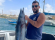 Sao-Vicente, Kapverden: Ein glücklicher Angler präsentiert stolz seinen beeindruckenden Fang, einen großen Wahoo, im Hafen. Die traumhafte Küstenkulisse und Segelboote im Hintergrund vervollständigen das maritime Erlebnis. Ideal für Angelliebhaber.