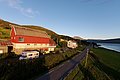 Malerische Landschaft in Nord-Norwegen: Ein rotes Bauernhaus, Wohnmobil und grüne Felder nahe einer Straße, die entlang eines ruhigen Fjords mit Bergblick verläuft, unter klarem, blauem Himmel.