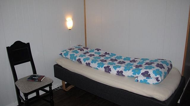 Ein gemütliches Einzelzimmer in Hitra, Norwegen: Bett mit bunter Blumenbettwäsche, Wandlampe, schwarzer Stuhl, ideale Unterkunft für Angelurlaub und Entspannung in skandinavischer Umgebung.