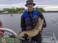 Eine Person hält einen großen Hecht auf einem Boot mit einer See- und Landkulisse im Hintergrund.