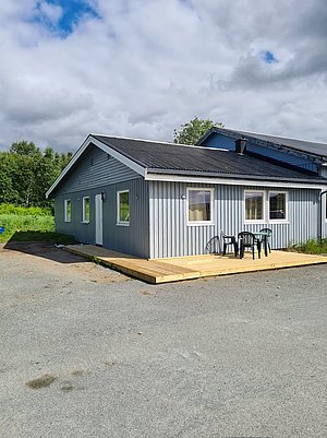 Gemütliches graues Ferienhaus in Nord-Norwegen. Vor dem Haus befindet sich eine Terrasse mit Tisch und Stühlen. Ruhige, grüne Umgebung ideal für einen entspannten Angelurlaub in der Natur. Wolkiger Himmel im Hintergrund.