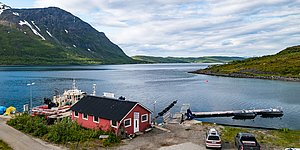 Die Aufnahme zeigt ein idyllisches Fischerdorf in Nord-Norwegen mit roten Hütten am Wasser, umgeben von majestätischen Bergen. Boote liegen am Pier, ideal für Angelreisen und Naturerlebnisse in Norwegens beeindruckender Fjordlandschaft.