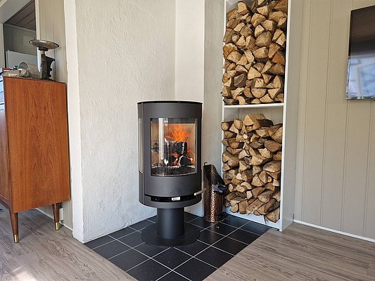 Ein gemütlicher Raum in Nordland, Norwegen. Moderner Kaminofen mit brennendem Feuer, neben einem Stapel Holzscheite. Perfekt für Angelurlaub-Abende. Warme, einladende Atmosphäre mit skandinavischem Charme.