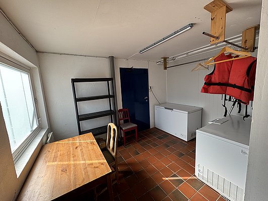 Ein praktischer Raum in Hitra, Norwegen, mit Tisch, Stühlen, Gefriertruhen und einem Regal. Ideal für Vorbereitung nach erfolgreichem Angelurlaub. Schwimmweste und Holzkleiderbügel für Sicherheit und Komfort.