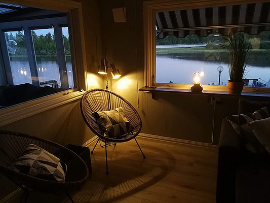 Gemütliches Wohnzimmer mit Rattan-Stühlen, indirekter Beleuchtung und Blick auf einen ruhigen See durch das Fenster.