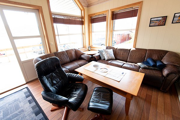 Gemütliches Wohnzimmer mit Ledersofas und Sesseln in einer Ferienhütte in Südwest-Norwegen. Helle Holzverkleidung und große Fenster bieten Blick ins Freie. Ideal für Angelreisen und Entspannung nach einem erfolgreichen Fang.